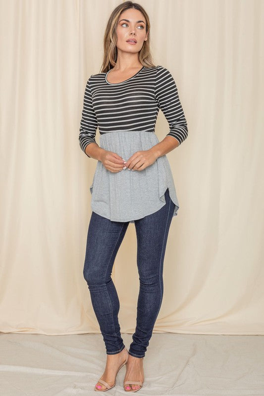 A woman elegantly stands in front of a beige curtain backdrop, wearing a stylish black striped top with a gray peplum and dark jeans, highlighting the sophistication of the Stripe Babydoll Tunic, proudly crafted in the United States.