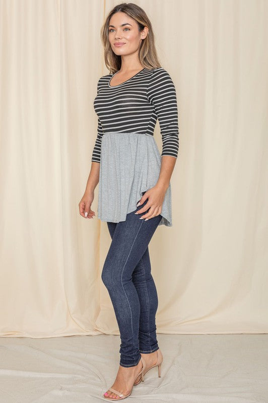 A woman wearing the Stripe Babydoll Tunic, designed with Rayon Spandex and featuring a gray flowy bottom, stands in front of a light curtain, gazing to the side.