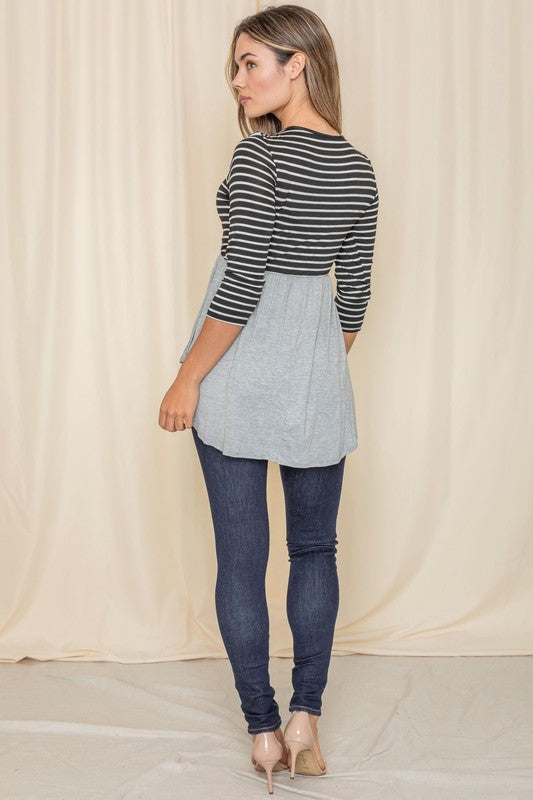 A woman elegantly stands in front of a beige curtain backdrop, wearing a stylish black striped top with a gray peplum and dark jeans, highlighting the sophistication of the Stripe Babydoll Tunic, proudly crafted in the United States.