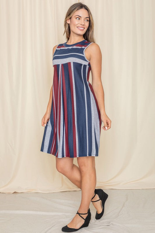 A person in a Sleeveless High Waist Mini Dress with vertical stripes of red, blue, and gray stands smiling while looking to the side in front of a light curtain backdrop.