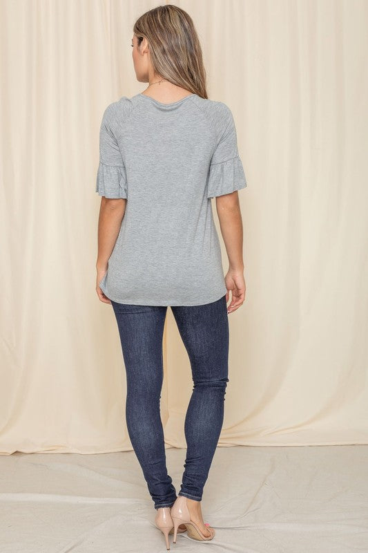 A woman in a Solid Ruffle Sleeve Top with a gray hue and sleeves accented with delicate ruffles smiles in front of a beige curtain, her look completed with a long necklace.