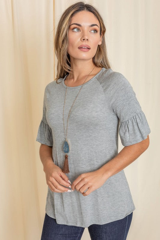 A woman in a Solid Ruffle Sleeve Top with a gray hue and sleeves accented with delicate ruffles smiles in front of a beige curtain, her look completed with a long necklace.