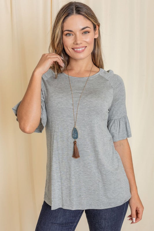 A woman in a Solid Ruffle Sleeve Top with a gray hue and sleeves accented with delicate ruffles smiles in front of a beige curtain, her look completed with a long necklace.