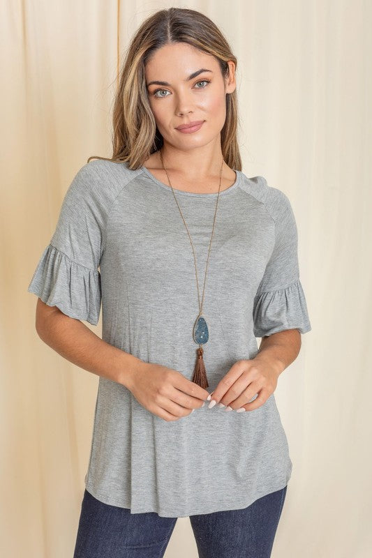 A woman in a Solid Ruffle Sleeve Top with a gray hue and sleeves accented with delicate ruffles smiles in front of a beige curtain, her look completed with a long necklace.