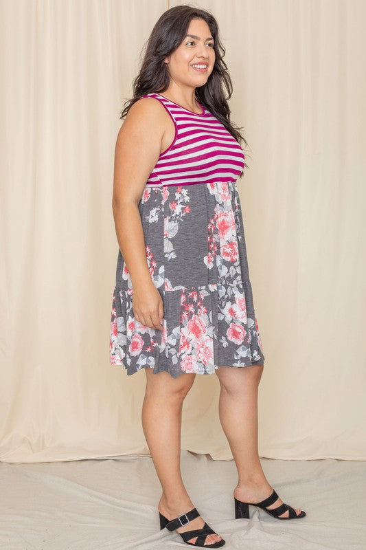 A woman in a Tiered Stripe Sleeveless Mini Dress, showcasing a striped magenta top and a floral gray skirt, stands against a beige background. Made in the United States, her outfit combines elegance with playful charm.