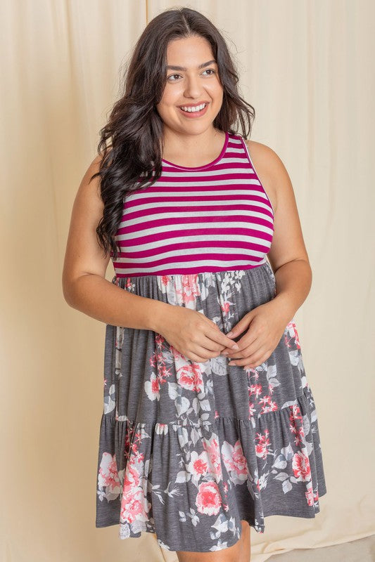 A woman in a Tiered Stripe Sleeveless Mini Dress, showcasing a striped magenta top and a floral gray skirt, stands against a beige background. Made in the United States, her outfit combines elegance with playful charm.