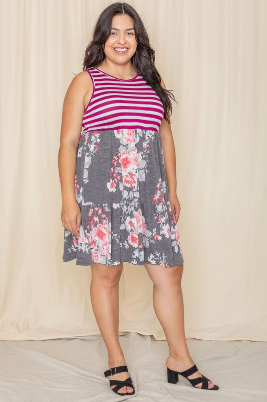 A woman in a Tiered Stripe Sleeveless Mini Dress, showcasing a striped magenta top and a floral gray skirt, stands against a beige background. Made in the United States, her outfit combines elegance with playful charm.