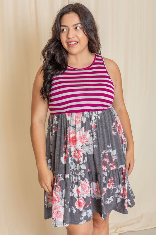 A woman in a Tiered Stripe Sleeveless Mini Dress, showcasing a striped magenta top and a floral gray skirt, stands against a beige background. Made in the United States, her outfit combines elegance with playful charm.