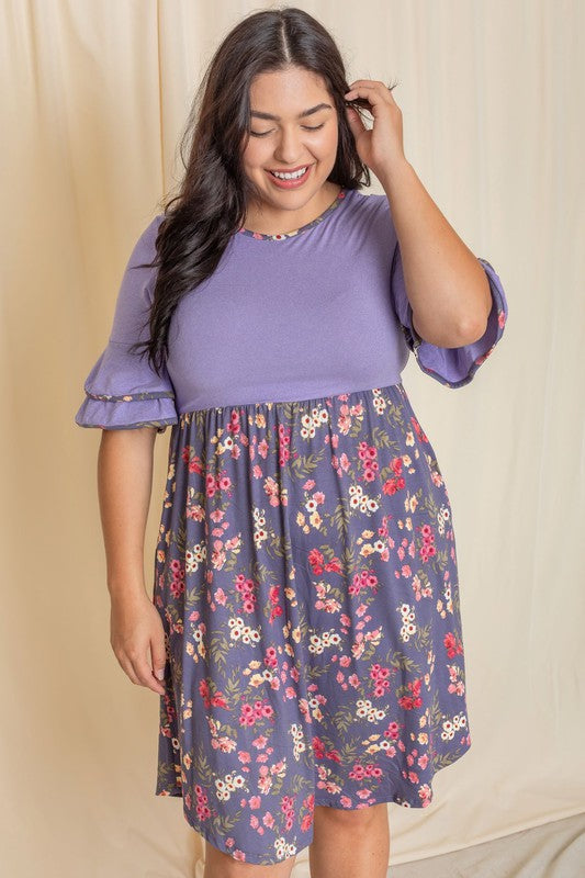 A woman gracefully poses in the Plus Double Ruffle Sleeve Midi Dress, showcasing a beautiful combination of a purple top and floral-patterned skirt in front of a cream curtain. The dress, crafted with care and proudly made in the United States, features elegant double ruffle sleeves.