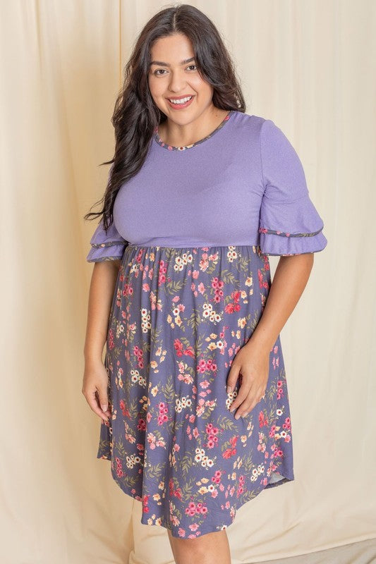 A woman gracefully poses in the Plus Double Ruffle Sleeve Midi Dress, showcasing a beautiful combination of a purple top and floral-patterned skirt in front of a cream curtain. The dress, crafted with care and proudly made in the United States, features elegant double ruffle sleeves.