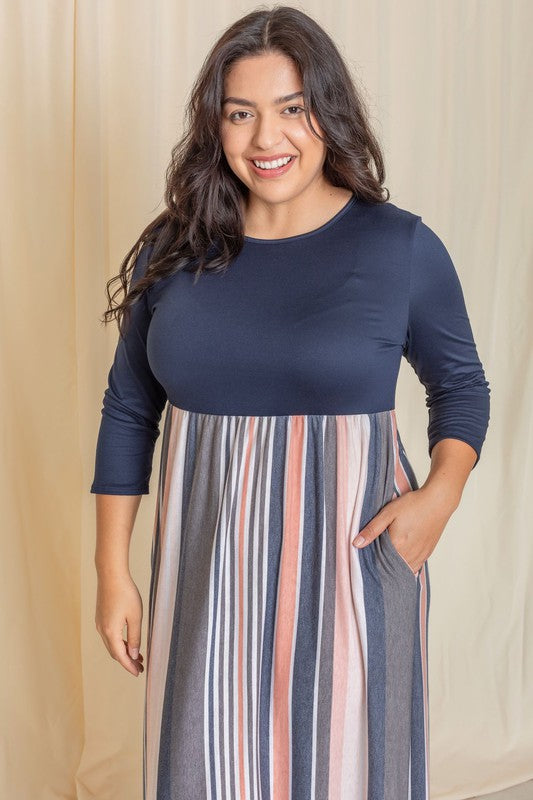 A person wearing the Plus Stripe Quarter Sleeve Maxi Dress, which showcases a navy top and a vertical striped skirt in blue, gray, and coral hues, stands in front of a beige curtain.