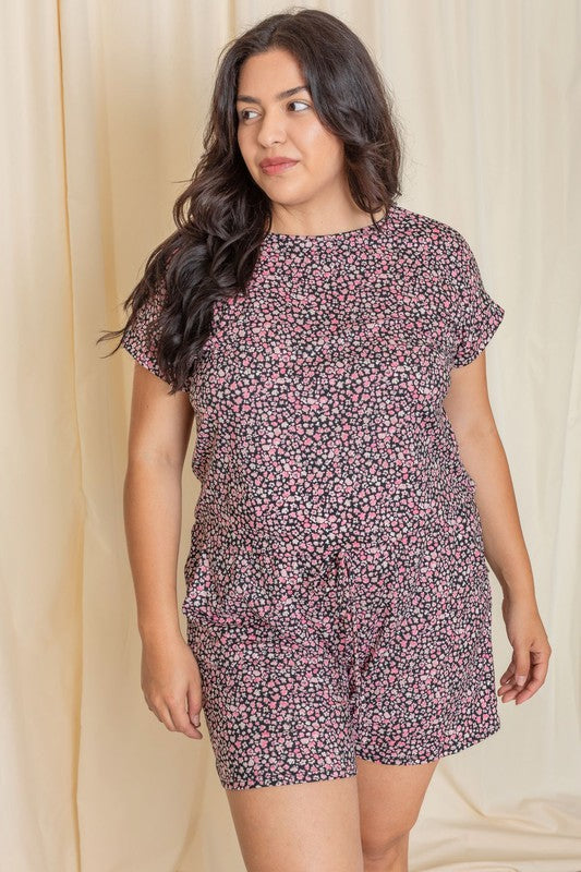 A woman wearing the Plus Short Sleeve Floral Print Romper, crafted from polyester-spandex, stands gracefully in front of a beige curtain.