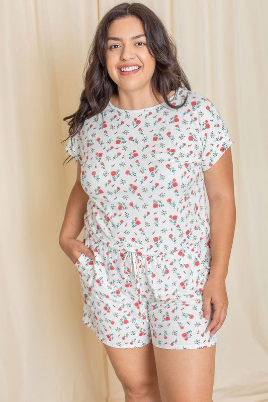 A woman wearing the Plus Short Sleeve Floral Print Romper, crafted from polyester-spandex, stands gracefully in front of a beige curtain.