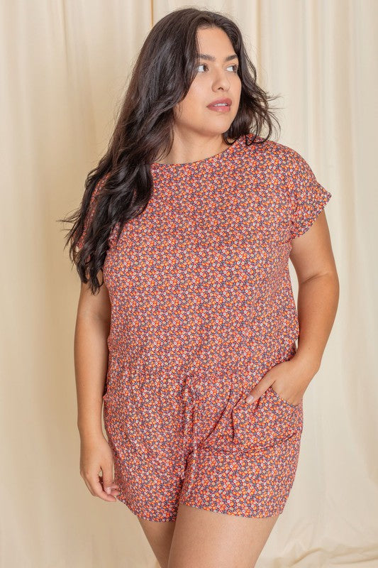 A woman wearing the Plus Short Sleeve Floral Print Romper, crafted from polyester-spandex, stands gracefully in front of a beige curtain.