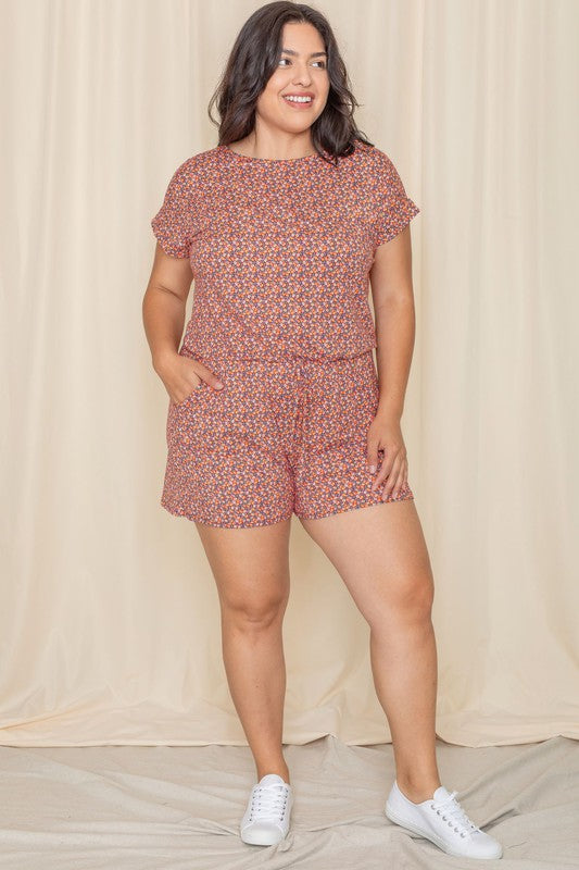 A woman wearing the Plus Short Sleeve Floral Print Romper, crafted from polyester-spandex, stands gracefully in front of a beige curtain.