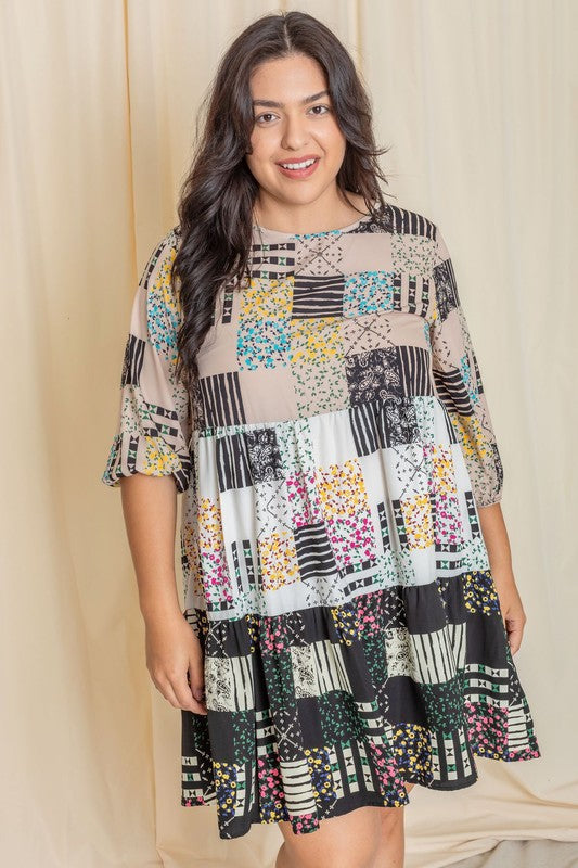 A woman elegantly poses against a beige curtain wearing the Plus Ditsy Floral Patch Work Dress, featuring a striking blend of black, white, and colorful designs crafted from woven polyester.