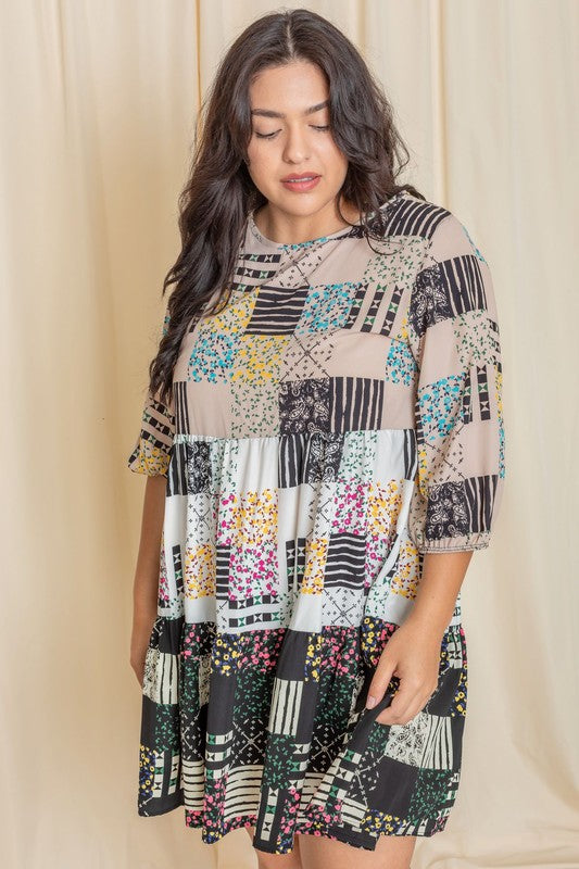 A woman elegantly poses against a beige curtain wearing the Plus Ditsy Floral Patch Work Dress, featuring a striking blend of black, white, and colorful designs crafted from woven polyester.