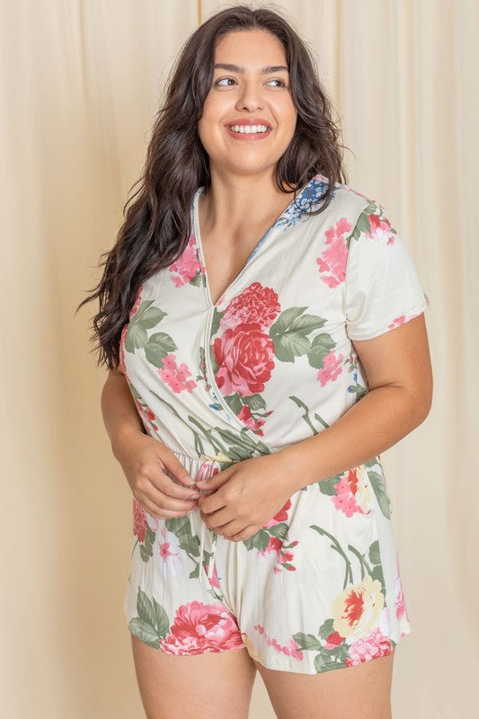 A person is standing in front of a beige curtain wearing the Plus Surplice Drawstring Floral Romper, which effortlessly blends comfort and elegance. This stylish outfit is proudly made in the United States.