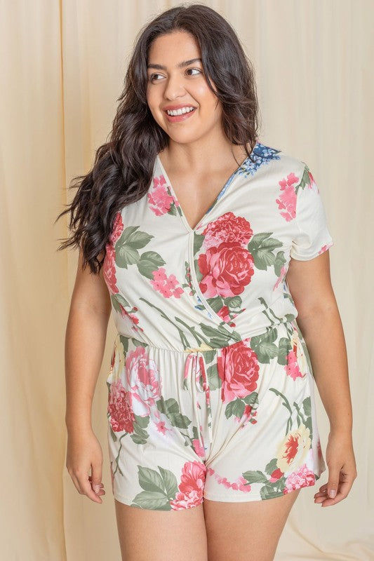 A person is standing in front of a beige curtain wearing the Plus Surplice Drawstring Floral Romper, which effortlessly blends comfort and elegance. This stylish outfit is proudly made in the United States.