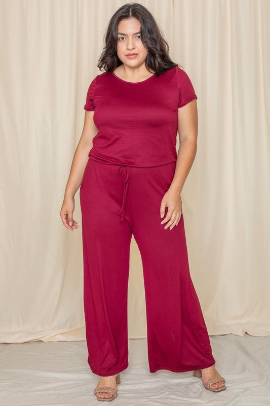 A person wearing the Short Sleeve Jumpsuit W/Pocket in dark gray, crafted in the United States, stands thoughtfully before a cream-colored curtain.