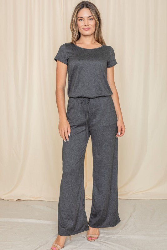 A person wearing the Short Sleeve Jumpsuit W/Pocket in dark gray, crafted in the United States, stands thoughtfully before a cream-colored curtain.