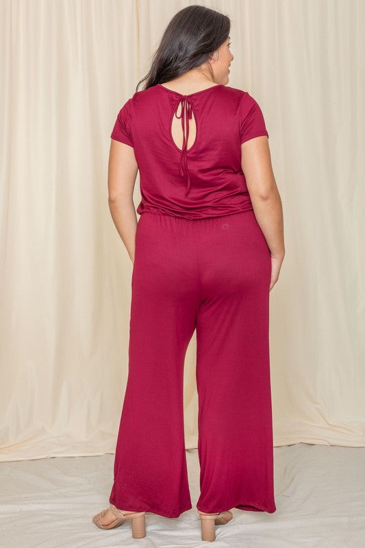 A person stands confidently in front of a beige curtain, showcasing a trendy Short Sleeve Jumpsuit W/Pocket.