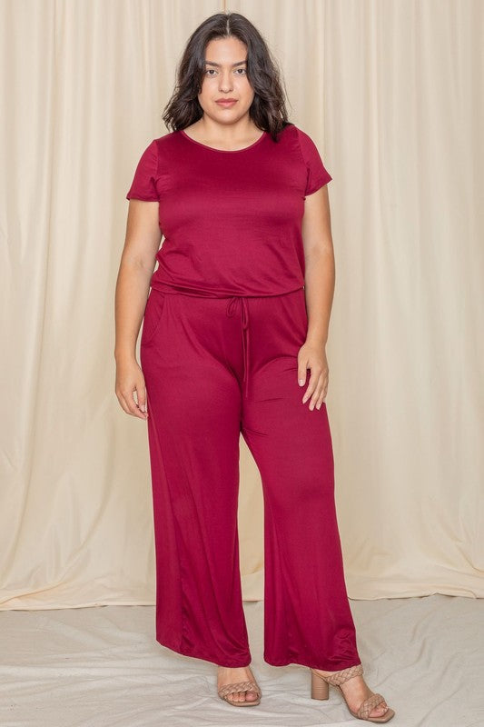 A person stands confidently in front of a beige curtain, showcasing a trendy Short Sleeve Jumpsuit W/Pocket.