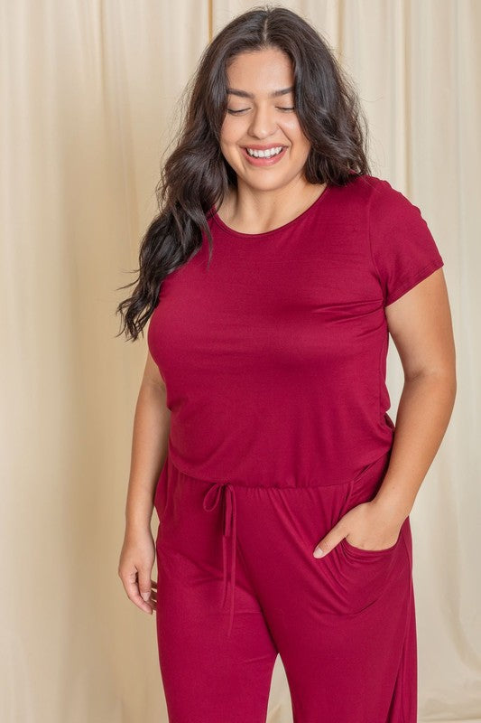 A person stands confidently in front of a beige curtain, showcasing a trendy Short Sleeve Jumpsuit W/Pocket.