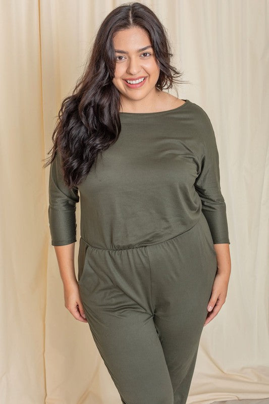 A woman wearing the Plus Quarter Sleeve Boat Neck Blouson Jumpsuit in green and sandals stands against a beige curtain backdrop.