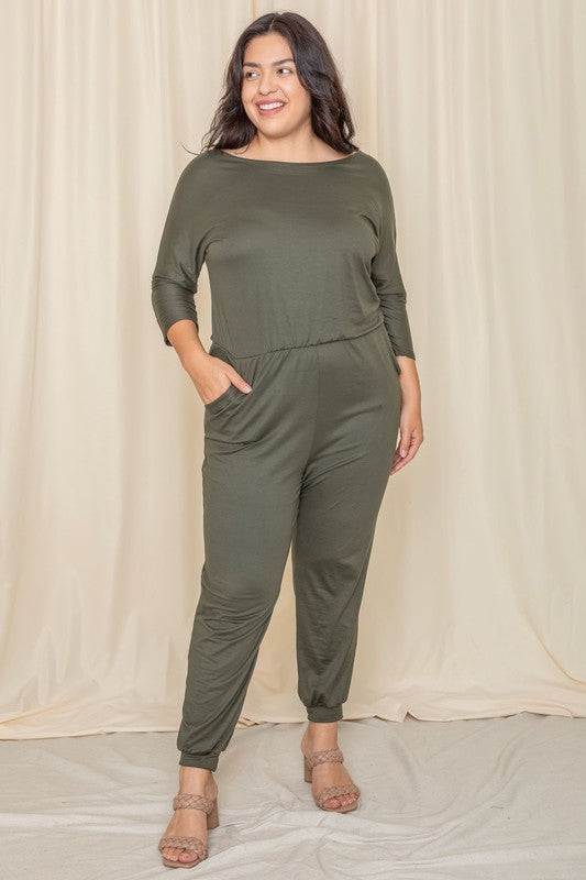 A woman wearing the Plus Quarter Sleeve Boat Neck Blouson Jumpsuit in green and sandals stands against a beige curtain backdrop.