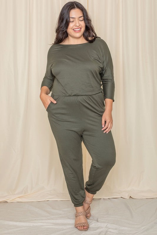 A woman wearing the Plus Quarter Sleeve Boat Neck Blouson Jumpsuit in green and sandals stands against a beige curtain backdrop.