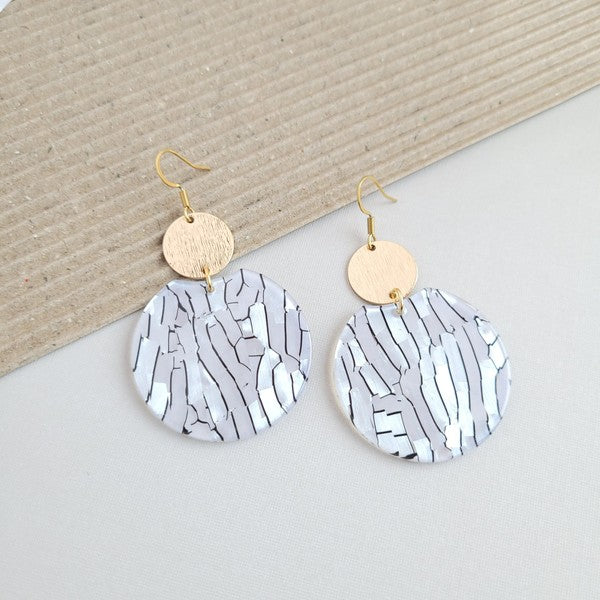 The Zoey Pearl earrings showcase a striking geometric design with gold-plated hooks. They feature a round gold disc paired with larger textured white circles that boast a shell-like finish, elegantly presented on a hexagonal marble surface.