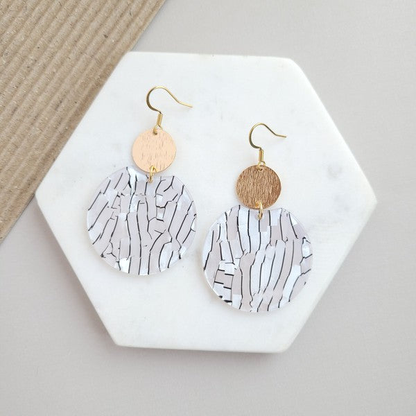 The Zoey Pearl earrings showcase a striking geometric design with gold-plated hooks. They feature a round gold disc paired with larger textured white circles that boast a shell-like finish, elegantly presented on a hexagonal marble surface.
