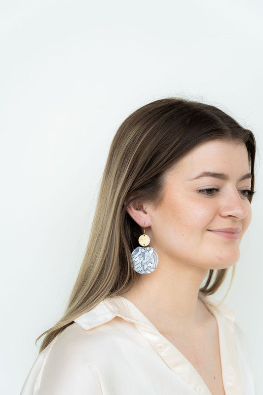 The Zoey Pearl earrings showcase a striking geometric design with gold-plated hooks. They feature a round gold disc paired with larger textured white circles that boast a shell-like finish, elegantly presented on a hexagonal marble surface.