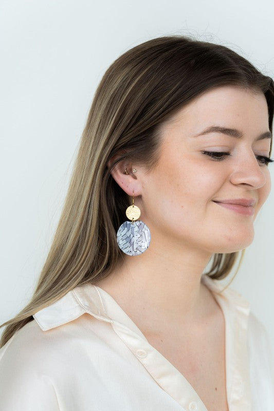 The Zoey Pearl earrings showcase a striking geometric design with gold-plated hooks. They feature a round gold disc paired with larger textured white circles that boast a shell-like finish, elegantly presented on a hexagonal marble surface.