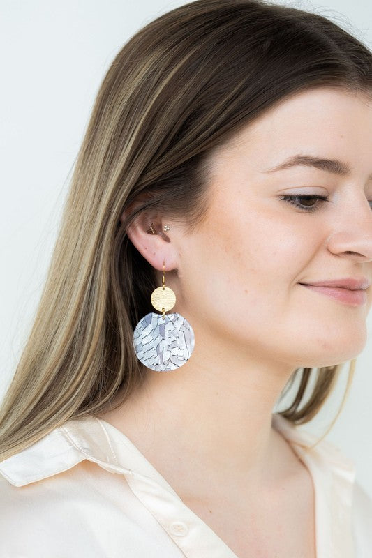The Zoey Pearl earrings showcase a striking geometric design with gold-plated hooks. They feature a round gold disc paired with larger textured white circles that boast a shell-like finish, elegantly presented on a hexagonal marble surface.