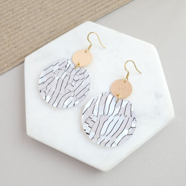 The Zoey Pearl earrings showcase a striking geometric design with gold-plated hooks. They feature a round gold disc paired with larger textured white circles that boast a shell-like finish, elegantly presented on a hexagonal marble surface.