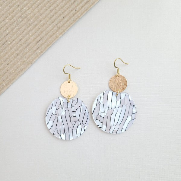 The Zoey Pearl earrings showcase a striking geometric design with gold-plated hooks. They feature a round gold disc paired with larger textured white circles that boast a shell-like finish, elegantly presented on a hexagonal marble surface.