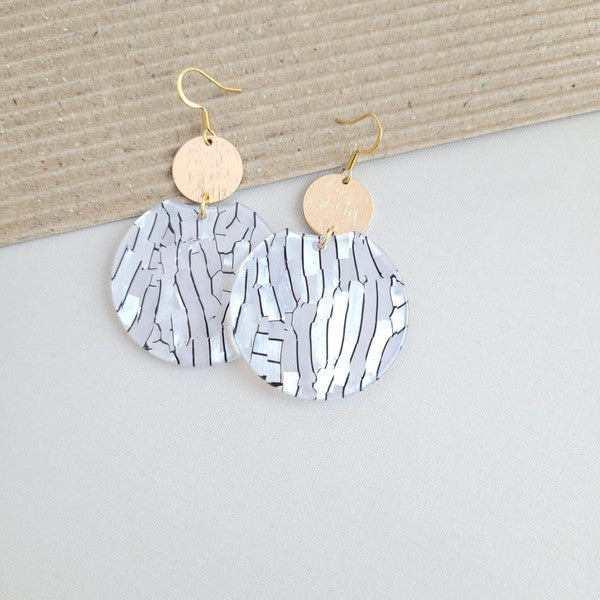 The Zoey Pearl earrings showcase a striking geometric design with gold-plated hooks. They feature a round gold disc paired with larger textured white circles that boast a shell-like finish, elegantly presented on a hexagonal marble surface.