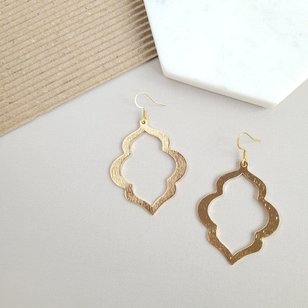 A person with long, wavy blonde hair is wearing the Talia Earrings in gold—a large geometric earring that serves as an elegant wardrobe staple, adding timeless charm to any look.