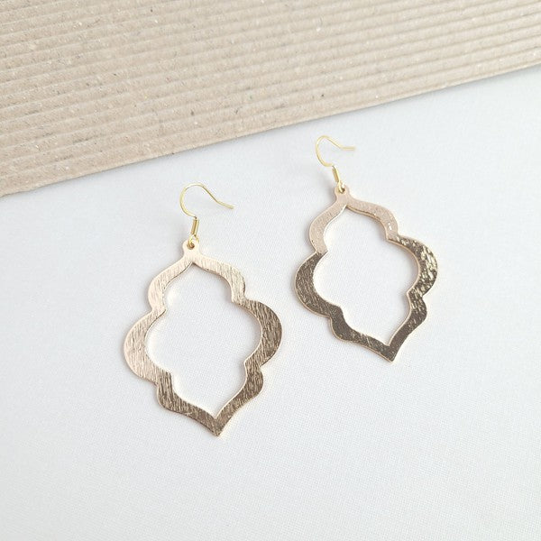 A person with long, wavy blonde hair is wearing the Talia Earrings in gold—a large geometric earring that serves as an elegant wardrobe staple, adding timeless charm to any look.
