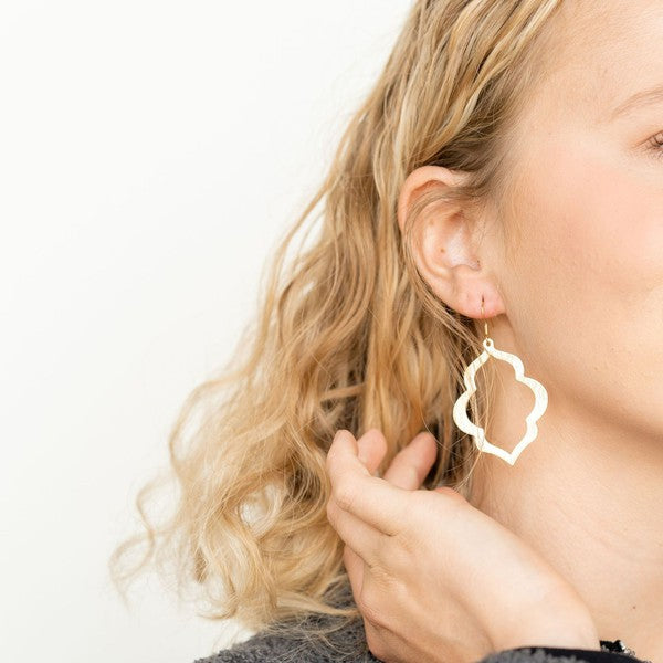 A person with long, wavy blonde hair is wearing the Talia Earrings in gold—a large geometric earring that serves as an elegant wardrobe staple, adding timeless charm to any look.
