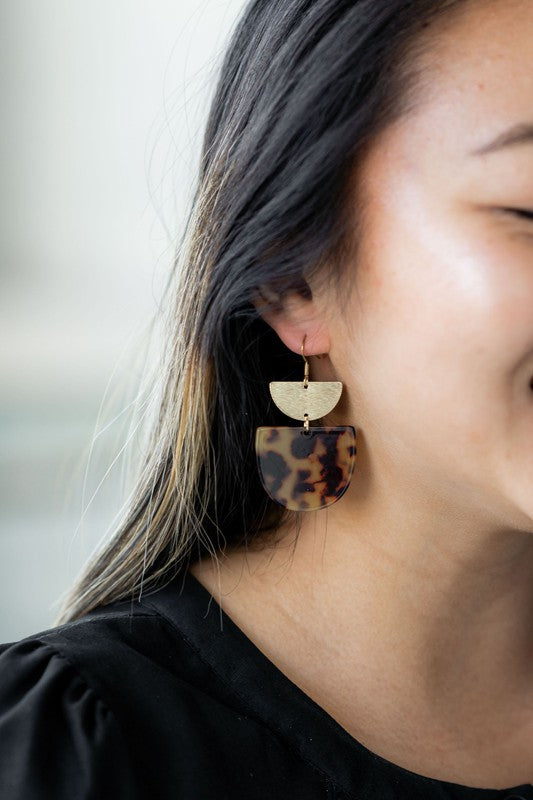 The Harper Pumpkin Spice Earrings make a bold statement with their gold-plated, geometric design elegantly displayed on a marble hexagonal platform.