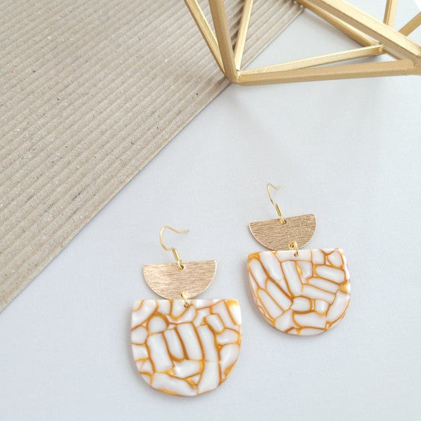 The Harper Pumpkin Spice Earrings make a bold statement with their gold-plated, geometric design elegantly displayed on a marble hexagonal platform.