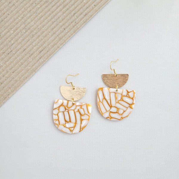 The Harper Pumpkin Spice Earrings make a bold statement with their gold-plated, geometric design elegantly displayed on a marble hexagonal platform.