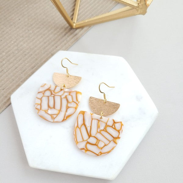 The Harper Pumpkin Spice Earrings make a bold statement with their gold-plated, geometric design elegantly displayed on a marble hexagonal platform.