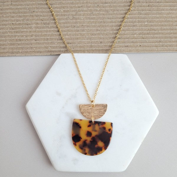 The Harper Tortoise Necklace showcases a gold-plated chain with a pendant featuring a small brushed gold semicircle and a larger tortoise acrylic semicircle, elegantly displayed on a hexagonal white slab.