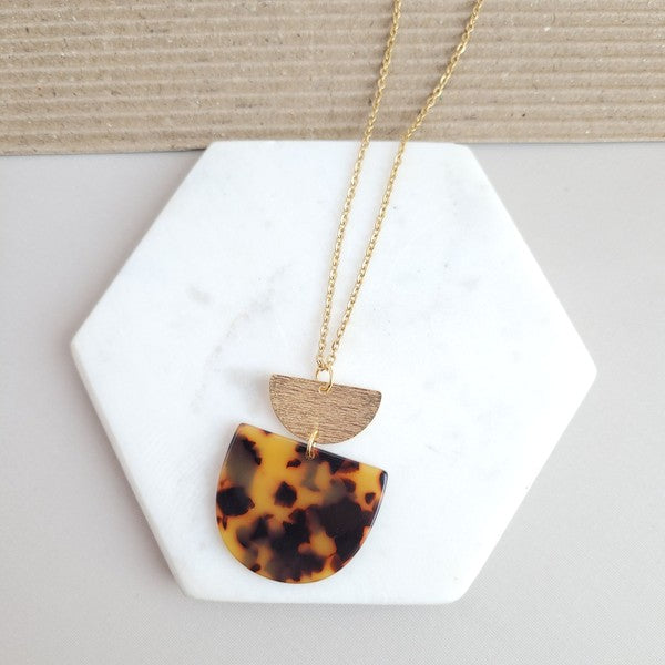 The Harper Tortoise Necklace showcases a gold-plated chain with a pendant featuring a small brushed gold semicircle and a larger tortoise acrylic semicircle, elegantly displayed on a hexagonal white slab.