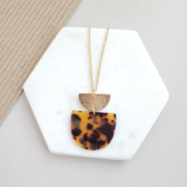 The Harper Tortoise Necklace showcases a gold-plated chain with a pendant featuring a small brushed gold semicircle and a larger tortoise acrylic semicircle, elegantly displayed on a hexagonal white slab.