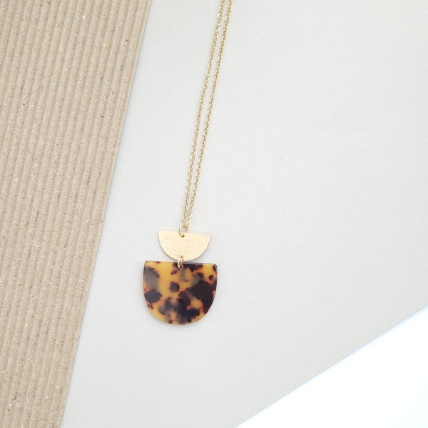 The Harper Tortoise Necklace showcases a gold-plated chain with a pendant featuring a small brushed gold semicircle and a larger tortoise acrylic semicircle, elegantly displayed on a hexagonal white slab.